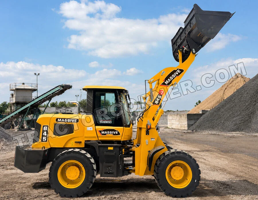 Yellow front-end loader at a quarry-style site, bucket raised high with a pile of gravel in the background, labeled 'MASSIVE' on the arm.